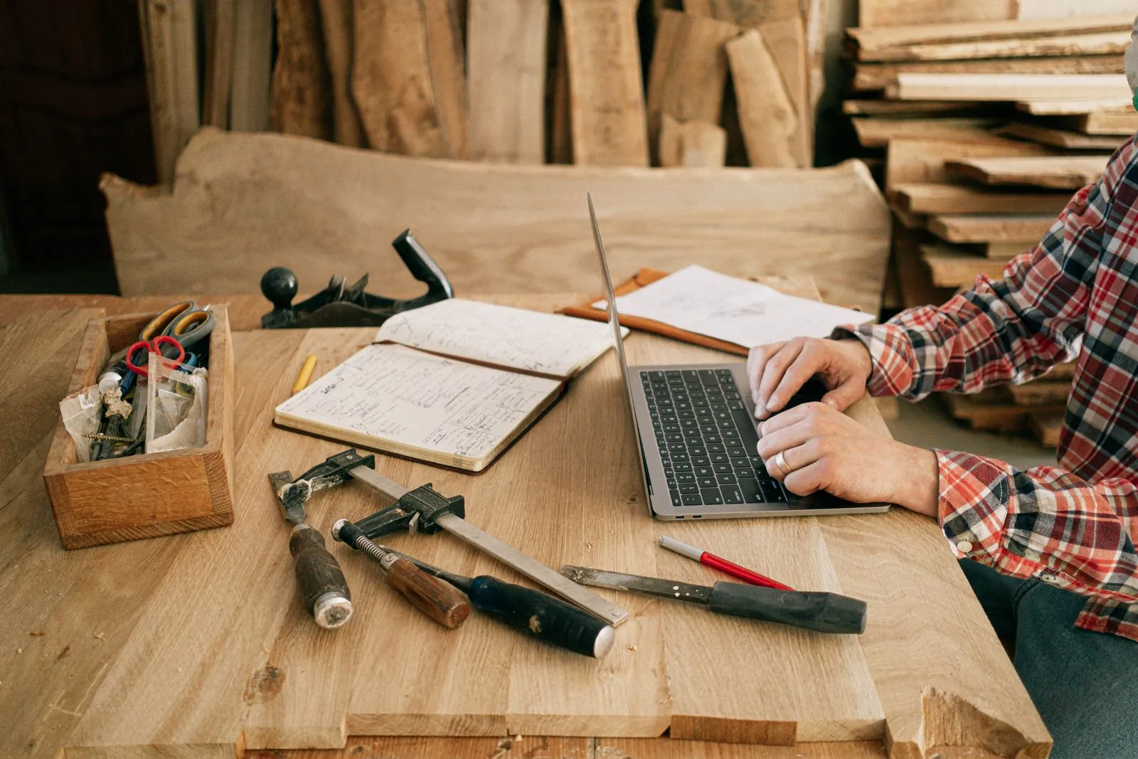 A carpenter works on a laptop at a workshop bench, beside a notebook and hand tools.