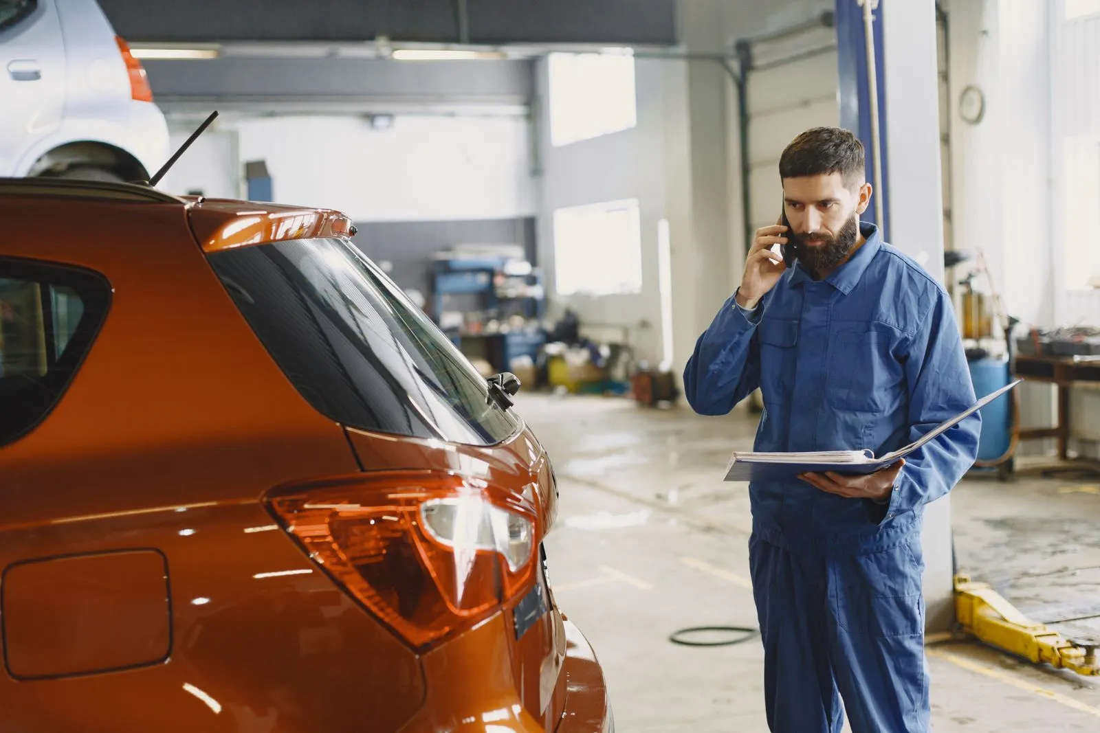 A mechanic in a workshop takes a phone call while holding a job folder.